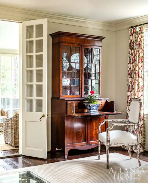 Living room with a dark wood secretary desk, a white upholstered chair with distressed frame, floral curtains, and a glass-top coffee table.