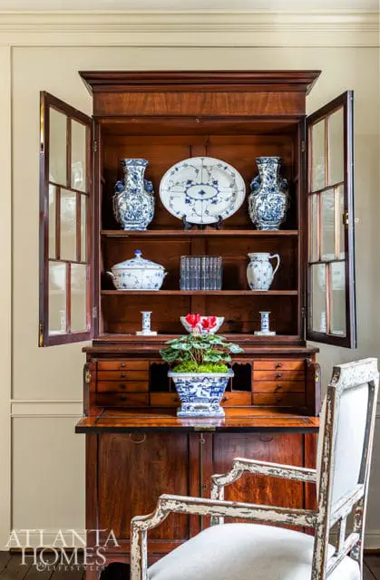 Antique wooden secretary desk with glass-fronted cabinet displaying blue and white porcelain vases, platters, and bowls. A potted plant sits on the desk, with a distressed white armchair in the foreground.