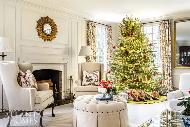 Living room with a decorated Christmas tree, fireplace, tufted ottoman, and wingback chairs. Floral curtains frame the windows.