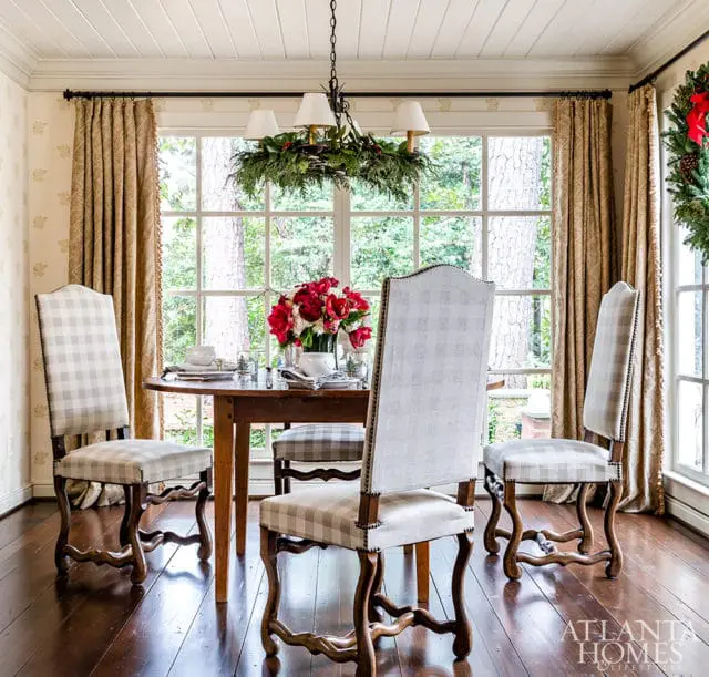 Dining room with a round wooden table set for a meal, surrounded by four upholstered chairs with a grey and white buffalo check pattern. A festive wreath hangs on the window.