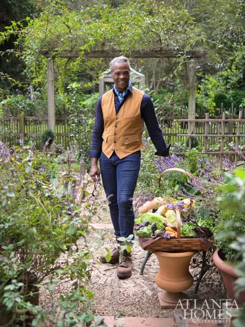 Man in a tan vest and blue scarf stands in a garden holding a basket of fresh vegetables.