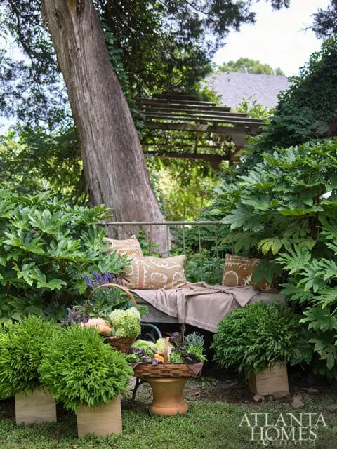 Garden bench with patterned pillows and a throw blanket surrounded by lush greenery, baskets of vegetables, and potted plants. A wooden pergola is visible in the background.