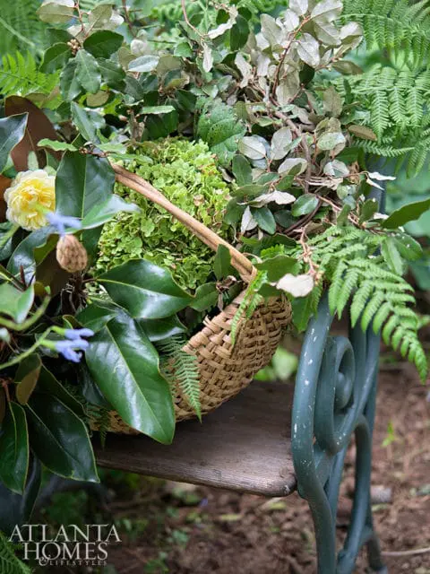 A woven basket overflowing with lush green foliage, ferns, and a yellow camellia sits on a rustic wooden bench with a dark green metal chair in the background.
