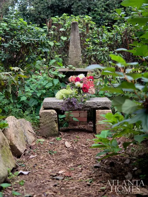 Stone garden bench with obelisk and floral arrangement surrounded by lush greenery and a dirt path. Atlanta Homes & Lifestyles logo in the corner.