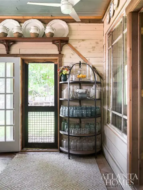 Rustic interior with white paneled walls, a blue ceiling, and a white ceiling fan. Features a wooden shelf with white plates and vases, a metal curio cabinet filled with glass jars, and a patterned rug.