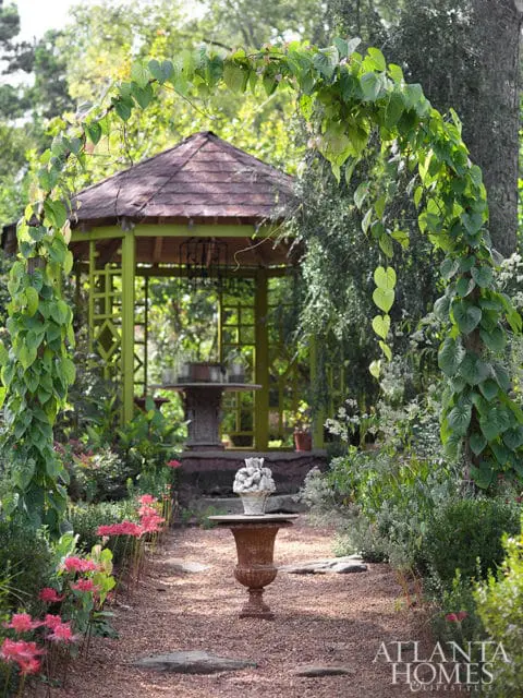 Garden path leading to a green gazebo, framed by a vine-covered archway and flanked by pink flowers and a weathered urn