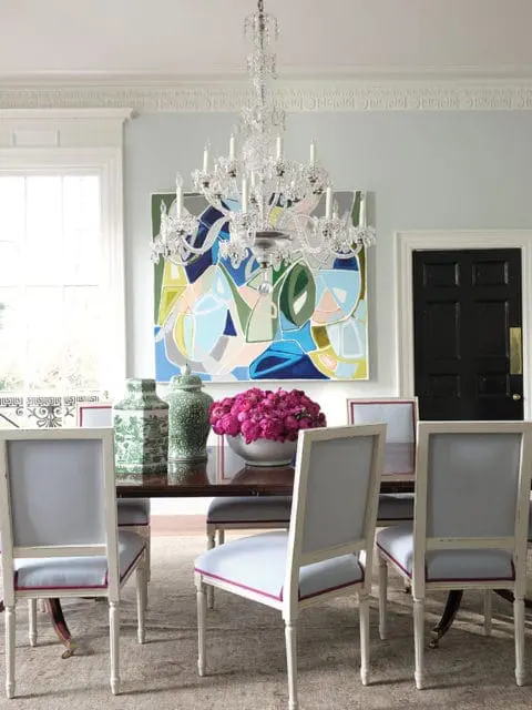Dining room with a crystal chandelier, abstract art, dark wood table, and upholstered chairs with light blue seats and dark pink trim.