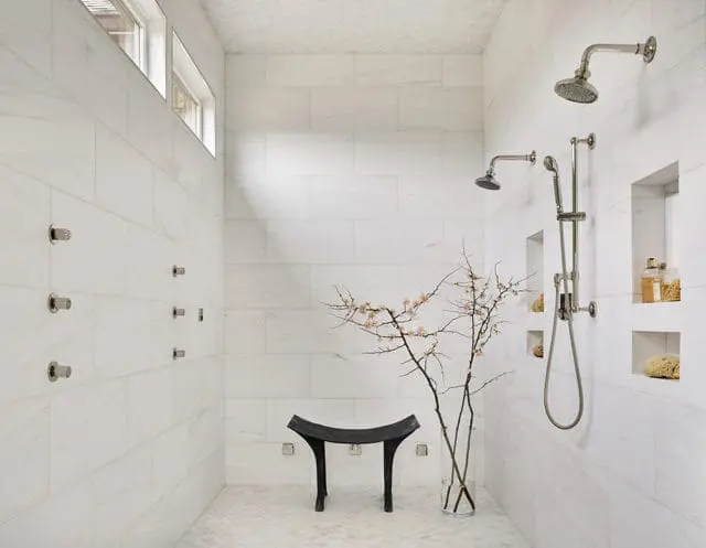 Master bathroom with white marble tile walls, multiple shower heads, recessed niches, a black bench, and a vase with branches and pink blossoms.