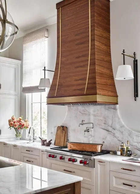 Kitchen with a large wooden range hood accented with brass trim, marble backsplash, and a professional gas range with red knobs.