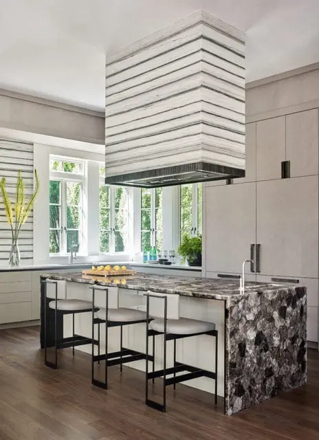Modern kitchen with a large island featuring a dark, multi-faceted stone countertop and a matching waterfall edge. A striped marble range hood hangs above the island, complemented by light wood cabinetry and white walls.