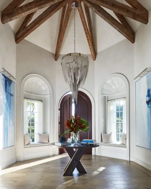 Entryway with vaulted ceiling, exposed wooden beams, a large chain-link chandelier, arched windows with built-in benches, and a round wooden table with books and a floral arrangement.