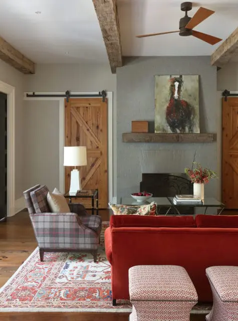 Living room with gray walls, exposed wooden beams, and a gray brick fireplace. Features include wooden barn doors, a plaid armchair, a red sofa, and a rug with red and blue patterns.