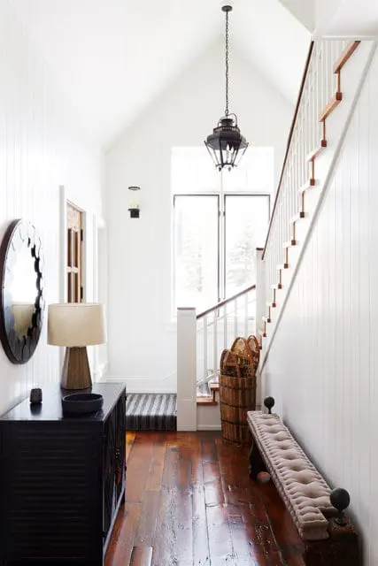 White hallway with vaulted ceiling, dark wood floors, a black lantern pendant light, a dark console table with a lamp and mirror, a tufted bench, and a staircase with white railings.