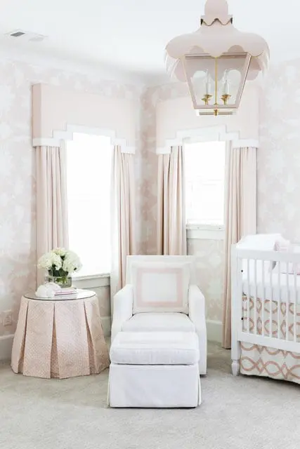 Nursery with pink floral wallpaper, white armchair and ottoman, a white crib with pink accents, and a pink pleated side table holding a vase of white hydrangeas.