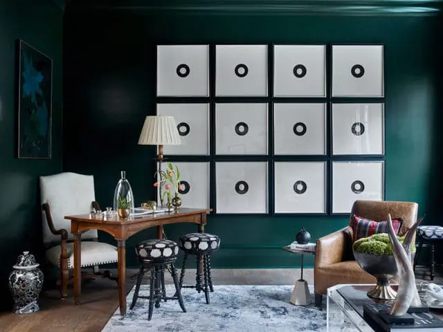 Dark green room with a wall of twelve framed prints of black circles on white backgrounds, a wooden desk with a lamp and decorative objects, a white upholstered chair, two patterned stools, and a leather armchair.