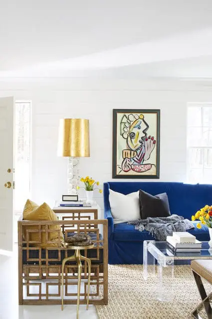 Living room with shiplap walls, a blue sofa with throw pillows, a wooden armchair with yellow cushions, and a colorful abstract painting.