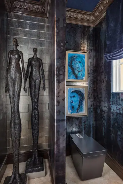 Dark bronze figurative sculptures stand against a wall of grey brick tiles. Two blue abstract portraits hang on a textured dark blue wall next to a modern dark grey toilet.