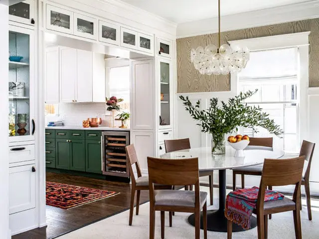 Dining room with a marble-top table and wooden chairs, adjacent to a kitchen with white and green cabinetry, subway tile backsplash, and a wine fridge.