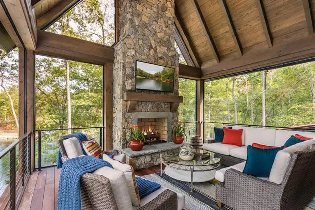 Covered porch with stone fireplace, white sectional sofa with teal and orange pillows, wicker chairs with blue throws, and a glass coffee table. Wood ceiling and walls with large windows overlook a wooded lake.