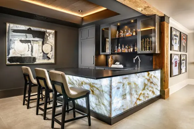 Modern bar area with a backlit onyx counter, dark gray cabinetry, a sink, and three upholstered bar stools. Abstract art hangs on the wall.
