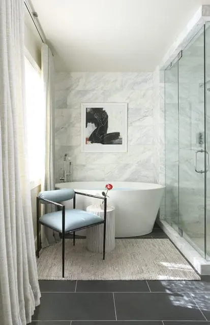 Master bathroom with a freestanding white tub, marble tile walls, glass-enclosed shower, and a light blue upholstered chair.