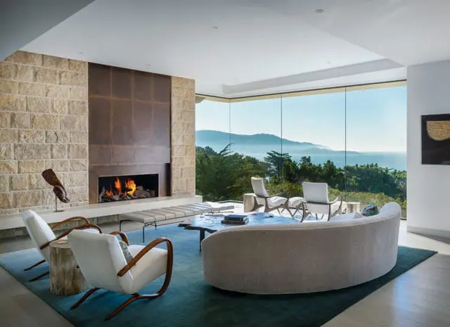 Living room with stone fireplace, curved sofa, white armchairs, and large windows overlooking the ocean and hills