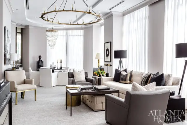 Modern living room with a cream sectional sofa, gray armchairs, a large circular chandelier, and floor-to-ceiling sheer curtains.