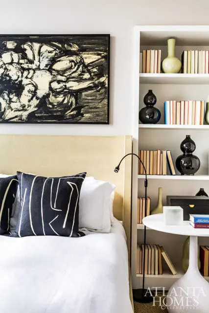 Bedroom with a light yellow upholstered headboard, black and white abstract art, and a white bookshelf filled with books and decorative vases.