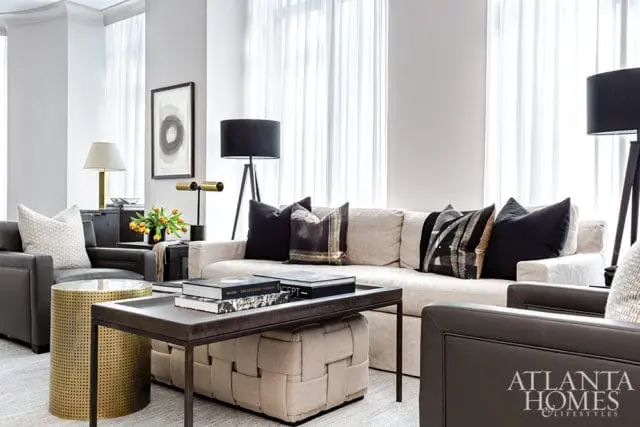 Living room with a neutral sectional sofa, dark leather armchairs, a metal coffee table with books, and a gold cylindrical side table.