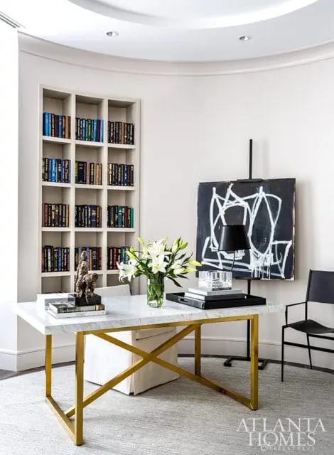 Home office with a marble desk featuring a gold X-frame base, a vase of white lilies, abstract art on an easel, and a tall bookshelf filled with books.