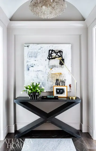 Entryway with a dark wood X-base console table topped with a vase of white tulips, a framed abstract artwork, and a brass task lamp. Above, a large crystal chandelier hangs from an arched ceiling.