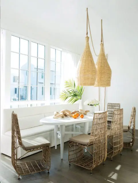 Bright dining room with a white oval table, white banquette seating, and four wicker chairs with tan cushions. Two large woven pendant lights hang above the table.