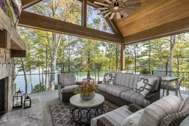 Covered patio with stone fireplace, sectional sofa, and dining set overlooking a lake surrounded by trees.