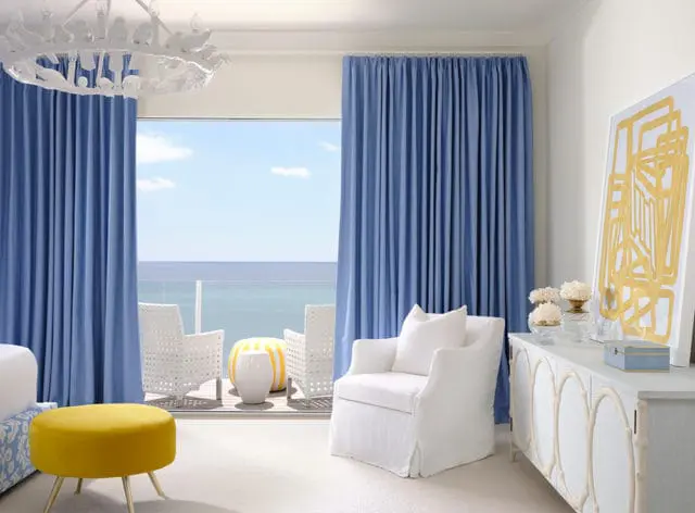 Bedroom with blue drapes, white slipcovered armchair, yellow ottoman, and a light blue dresser with oval inlays. A large window overlooks the ocean.