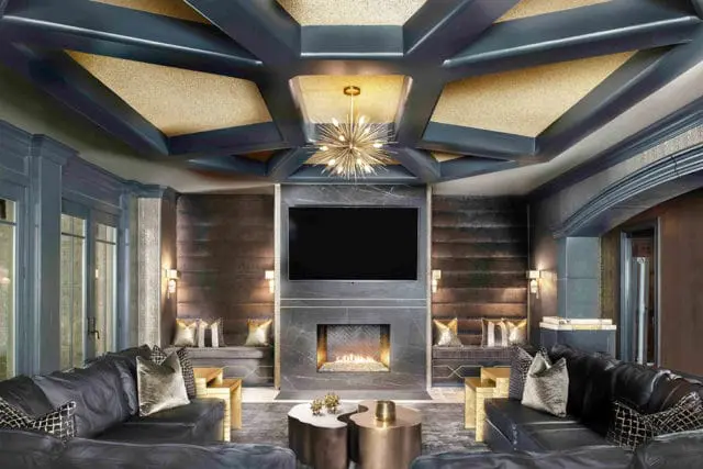 Dark gray sectional sofa facing a fireplace with a television above. The room features a coffered ceiling with gold accents, dark upholstered walls, and gold throw pillows.