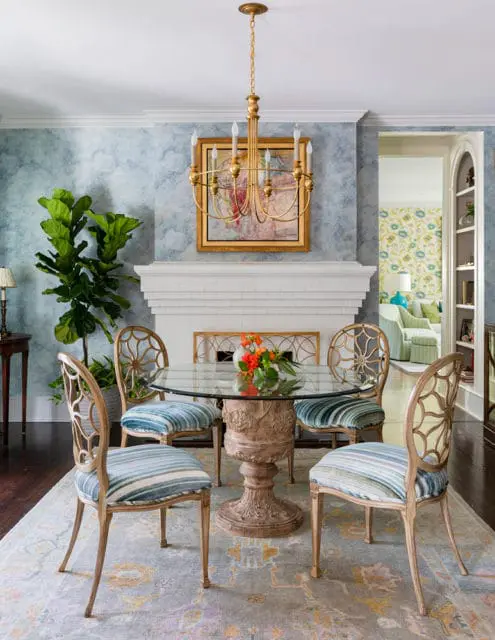 Dining room with a glass-top table, ornate pedestal base, and four chairs with striped blue upholstery. A white fireplace with a gold chandelier hangs above.