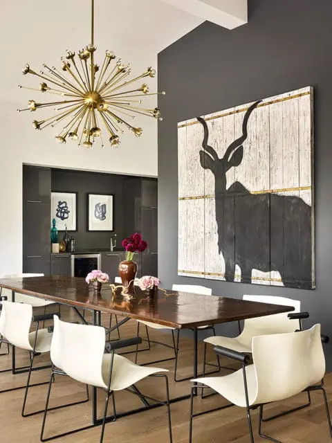 Dining room with dark wood table, cream chairs with black arms, sputnik chandelier, and large artwork of a kudu silhouette on a dark gray wall.