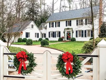 White colonial-style house with black shutters and a detached garage, decorated for Christmas with wreaths and garlands.