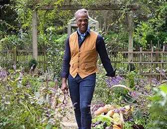 Man in a tan vest and blue shirt walks through a garden carrying a basket of vegetables.