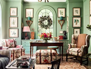 Green room with floral upholstered chairs, a dark wood desk, and botanical prints on the walls. A large arched window features a wreath.
