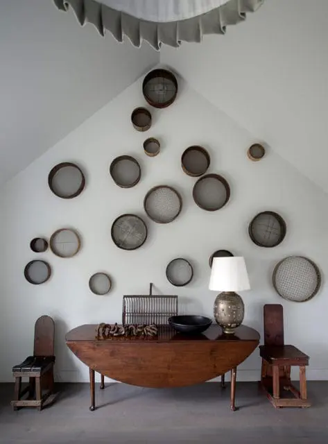 Wall decorated with a collection of vintage sieves and strainers of various sizes, arranged above a wooden drop-leaf table holding a lamp, bowl, and decorative cage.