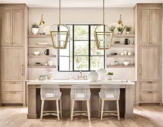 Light wood kitchen with a large island featuring a white countertop and three woven counter stools. Two brass pendant lights hang above the island, with open shelving and a window in the background.