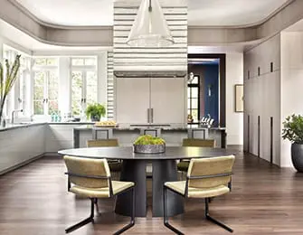 Modern kitchen with a large oval dining table and four chairs in the foreground, a central island with a range hood, and light gray cabinetry.
