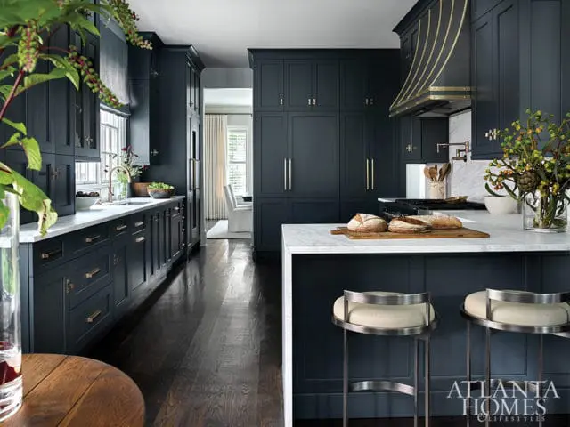 Dark blue kitchen with marble countertops, brass hardware, and a decorative range hood. Two bar stools with cream upholstery are at the island.