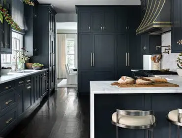Dark blue kitchen with white marble countertops, brass hardware, and a decorative brass range hood. A kitchen island with bar stools is in the foreground.