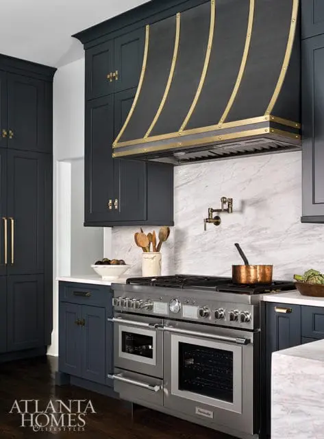 Dark blue kitchen cabinets with brass hardware, a stainless steel range with a copper pot, and a black and brass range hood over white marble backsplash.