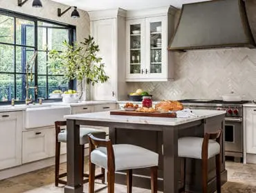 Cream kitchen with marble island, dark wood base cabinets, white marble countertops, white apron sink, brass faucet, and large black-framed window overlooking greenery.