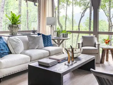 Living room with white sectional sofa, dark wood coffee table, and large windows overlooking a forest.