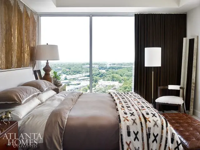 Bedroom with large windows overlooking a city skyline, featuring a king-sized bed with brown and white bedding, a decorative throw blanket, and a leather ottoman.