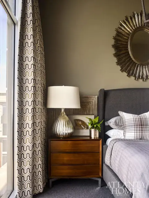 Bedroom with a dark gray upholstered headboard, gray bedding, and a patterned throw pillow. A wooden nightstand holds a silver lamp and a small plant. A sunburst mirror hangs on the olive green wall.